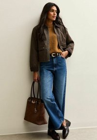 Vrouw in een bruine cropped jacket, tan sweater, blauwe wijde jeans, donkere loafers, met een grote bruine handtas, leunend tegen een beige muur.