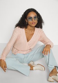 Blusa envolvente texturizada rosa claro com mangas longas, combinada com jeans flare azul claro e mules de salto bloco creme. Óculos de sol oversized transparentes.