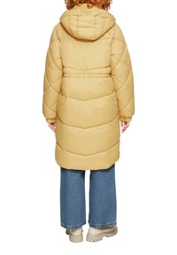 Lang, gele pufferjas met een capuchon, voorzien van een gestikt ontwerp en elastische manchetten, gedragen over flared blauwe jeans en beige schoeisel.