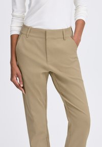 Bež hlače s krojem, izdelane iz gladkega materiala, z obstranskimi žepi in standardnim pasom. Oblikovane za ozek kroj.
