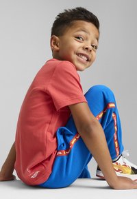 Camiseta roja de manga corta combinada con pantalones de chándal azules que tienen franjas laterales rojas y amarillas. Lleva zapatillas multicolores.