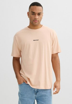 Hombre vistiendo una camiseta color melocotón con la inscripción "Miami Beach" y jeans azul claro, de pie con una mano en el bolsillo, sobre un fondo liso.