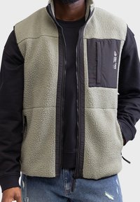 Man in een lichtgrijze fleecevest met een zwarte borstzak, over een zwart langerm T-shirt en gescheurde blauwe jeans, met zijn handen in de vestzakken.