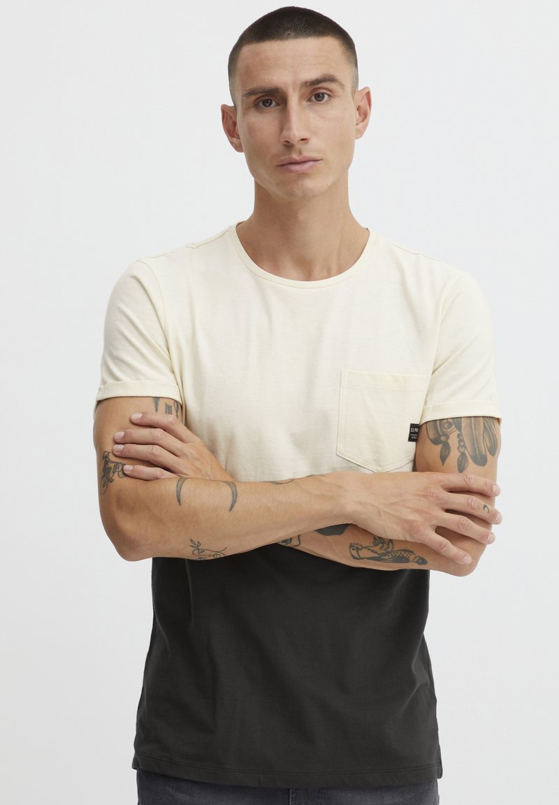 T-shirt med korte ærmer, der har en lys cremefarvet øverste del og en ensfarvet sort nederste del. Den har en brystlomme og en rund halsudskæring.