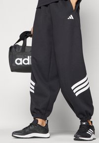 Pantaloni da allenamento neri con accenti a strisce bianche sui lati, abbinati a scarpe da ginnastica nere e a una borsa sportiva nera coordinata.