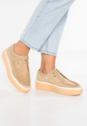 Beige Wildleder-Sneaker mit abgerundetem Zeh, Schnürdesign und einer klobigen hellorangen Sohle mit weißem Akzent.