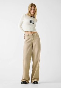 Pull court en maille crème avec le numéro 81, associé à un pantalon beige à jambes larges. Le pull présente un motif texturé, tandis que le pantalon est en tissu lisse.