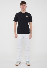 T-shirt in cotone nero con collo rotondo, caratterizzato da un piccolo logo colorato sul lato sinistro del petto. Abbinato a jeans di denim bianchi e sneakers di colore chiaro.