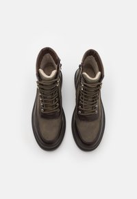 Botas de cuero marrón hasta el tobillo con un acabado texturizado, cuello acolchado, ojales de metal y cordones. Forro suave visible en la parte superior.