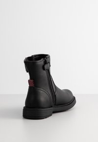 Botte noire en cuir avec fermeture éclair latérale, languette de tirage en tissu rouge au talon et semelle plate rainurée sur fond blanc.