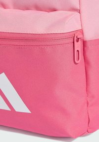 Detailný záber na ružovú látkovú kapsu batohu s ružovým zipsom a časťou bieleho geometrického loga v dolnom ľavom rohu.