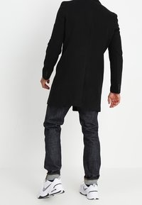 Zwarte lange jas van wol, met een geknipt kraag en een getailleerde pasvorm. Draagt over donkere denim jeans en witte sneakers.