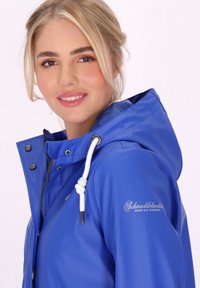 Kék vízálló dzseki kapucnival, patentgombokkal és húzózárral. Logo a tenyéren, sima textúra és modern dizájn.
