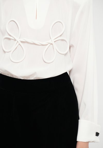 White blouse with decorative twisted loops at the chest and black velvet pants. Blouse has long sleeves with a contrasting cuff.