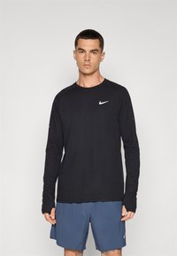 Chemise noire à manches longues avec un col rond et un logo Nike blanc, associée à un short bleu. Le tissu semble texturé et léger.