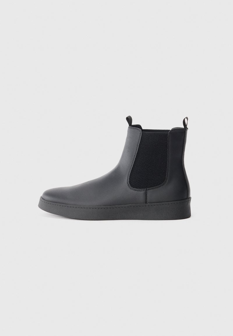 Botas Chelsea negras de material sintético liso, con paneles laterales de malla, lengüetas de agarre y suela de goma plana.
