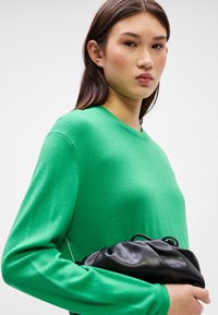 Πράσινο πλεκτό πουλόβερ με μακριά μανίκια και στρογγυλή λαιμόκοψη. Το μοντέλο κρατά μια μαύρη δερμάτινη τσάντα φακέλου. Λείο υλικό, απλός σχεδιασμός.