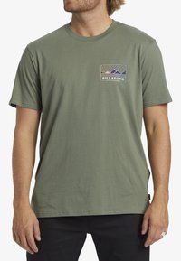 Short-sleeve green cotton t-shirt featuring a rectangular graphic with mountains and rays in yellow, blue, and white, branded "Billabong."