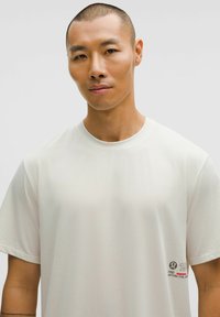 Homme asiatique avec les cheveux courts portant un t-shirt de sport blanc avec un petit logo et du texte en bas à droite.