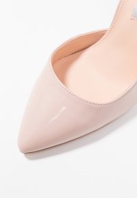 Lesklé světle růžové lodičky s špičatou špičkou, hladkým povrchem a viditelným šitím podél obvodu na bílém pozadí.