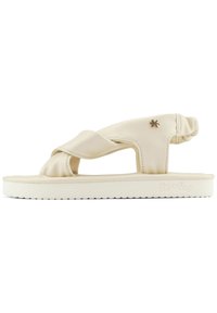 Beige flip-flop sandal with a cross strap design, made of soft material, featuring a ribbed sole and a small metallic accent.