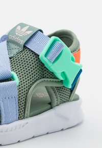 Zelené síťové sandály s světle modrými a oranžovými akcenty, plastovou sponou a bílou podrážkou; mají otvory pro větrání a flexibilní design.