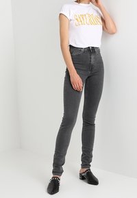 Camiseta blanca con letras amarillas, jeans ajustados de tiro alto grises y mule negros con tachuelas blancas. Textura de tela suave, diseño ajustado.