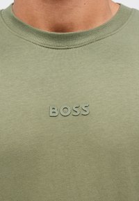 Õlivärvi roheline pusa, tehtud pehmest kangast; sellel on tekstuuriga ümara kaelusega lõige ja rinnal kõrgendatud "BOSS" logo.