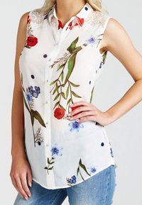 Blouse sans manches à motifs floraux avec une base blanche, présentant des fleurs rouges et bleues, des feuilles vertes, une finition en dentelle sur les épaules et une fermeture à boutons sur le devant.