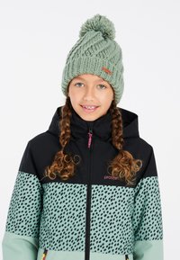 Berretto lavorato a maglia verde con pom-pom, indossato con una giacca nera e teal con un motivo a pois e una cerniera con una linguetta rosa.