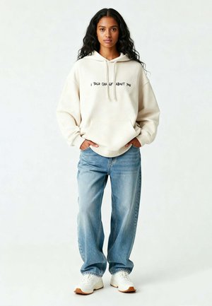 Felpa crema con testo nero, tasca frontale, polsini a coste; abbinata a jeans azzurri chiari e sneakers bianche con suole marroni.