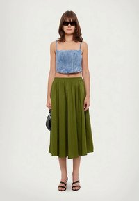 Top scurt din denim cu bretele subțiri, asortat cu o fustă lungă plisată verde. Sandale negre și o geantă mică neagră completează look-ul.