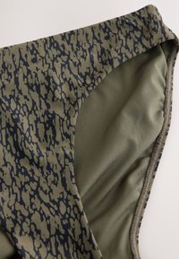 Gros plan du bas de bikini vert olive avec des taches abstraites irrégulières bleu marine foncé et une doublure en tissu lisse à l'intérieur.