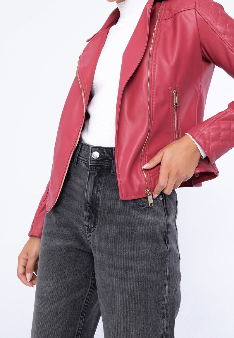Červená kožená motocyklová bunda s prošívanými detaily na ramenou, stříbrnými zipy a černými džínami s vysokým pasem, které mají poničenou texturu a klasické knoflíky.