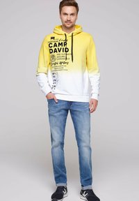 Sudadera con degradado de amarillo a blanco, con texto gráfico, bolsillo frontal tipo canguro, capucha con cordones, combinada con jeans de color azul claro y zapatillas negras.