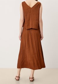 Brown pleated sleeveless top with V-neck and matching long skirt. Flowing fabric creates a textured appearance. Flat sandals visible.