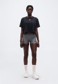 Γυναίκα που φοράει μαύρο κροπ τοπ και γκρι σορτς κρατάει αλτήρες 1 κιλού όρθια μπροστά σε απλό λευκό φόντο.