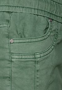 Tejido de mezclilla verde con un acabado texturizado, que presenta un bolsillo frontal con herrajes de metal y detalles de costura en contraste.