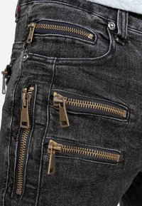 Close-up of black denim pants with four brass-colored horizontal and vertical zippers on pocket areas.