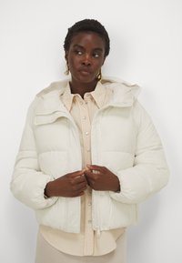 Giacca a vento leggera color crema, design trapuntato, dotata di cappuccio rimovibile e chiusura con zip, abbinata a una camicia beige con bottoni sotto.