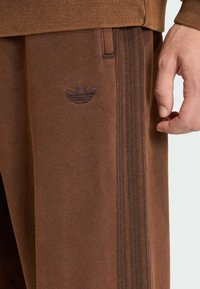 Braune Sporthose aus strukturiertem Stoff, mit einem tonal gehaltenen Logo und drei vertikalen Streifen in einem dunkleren Farbton. Hand liegt auf dem Bund.