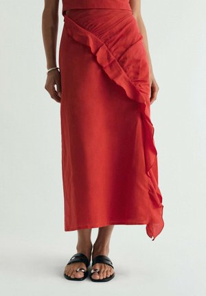 Mujer con una falda larga roja con un detalle diagonal con volantes, combinada con sandalias negras de punta abierta, de pie sobre un fondo liso.