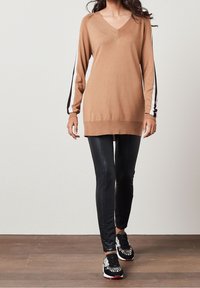 Brauner Strickpullover mit V-Ausschnitt und langen Ärmeln, die mit schwarz-weißen Streifen gestaltet sind. Kombiniert mit schwarzen Leggings und gemusterten Sneakern.
