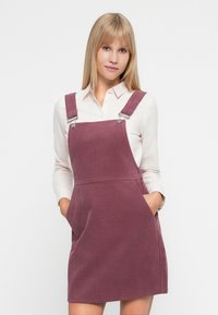 Vestido de tirantes de pana en rosa empolvado, con tirantes ajustables, bolsillos delanteros y un acabado texturizado. Combinado con una camisa blanca de botones.