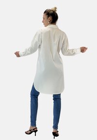 Camicia lunga bianca con colletto e polsini a bottoni, realizzata in tessuto leggero; abbinata a jeans skinny blu e tacchi neri.