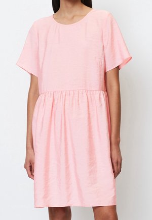 Femme portant une robe rose clair ample à manches courtes avec un col rond et un détail froncé à la taille sur un fond uni.