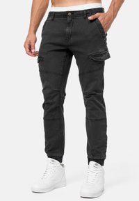 Pantalon cargo noir avec poignets élastiques, fermeture à boutons, poches latérales et détails cousus. Porté avec des baskets montantes blanches.