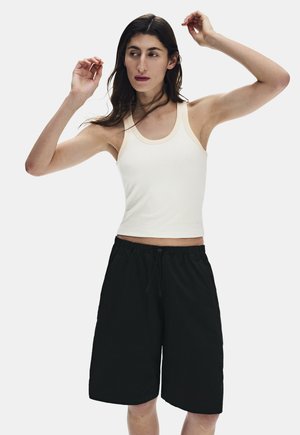 Femme aux longs cheveux foncés portant un débardeur blanc et un short noir ample arrivant au genou, posant les bras levés devant un fond uni.
