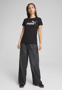 Melna kokvilnas T-kreklu ar balto "PUMA" logo, kombinēta ar brīvām tumšām džinsu biksēm un melniem apaviem ar struktūru zoli.