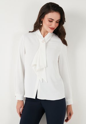 White blouse with a bow tie collar, long sleeves, and button closure. Features a pearl accent near the collar and has a smooth texture.